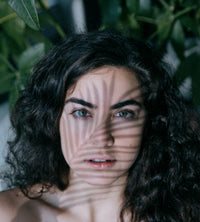 Woman with wavy hair and green eyes, with shadows cast across her face against a blurred natural background.