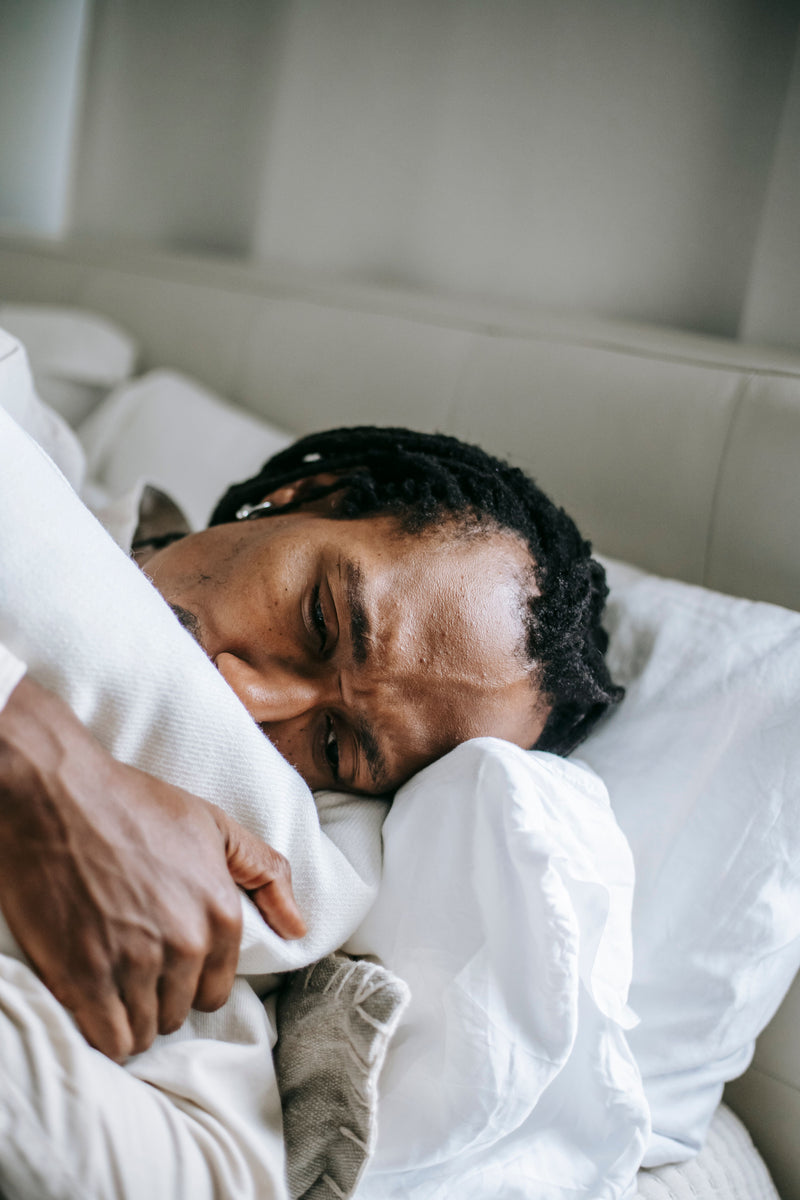 A close-up lifestyle shot of a man sleeping soundly in bed, wrapped in a white blanket, with soft neutral tones from the headboard and pillows in the background.