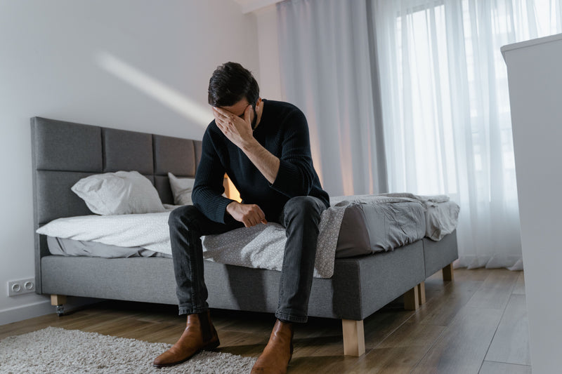 A man in a black long-sleeve shirt and dark jeans sits dejectedly on a gray upholstered bed in a bright, modern bedroom.