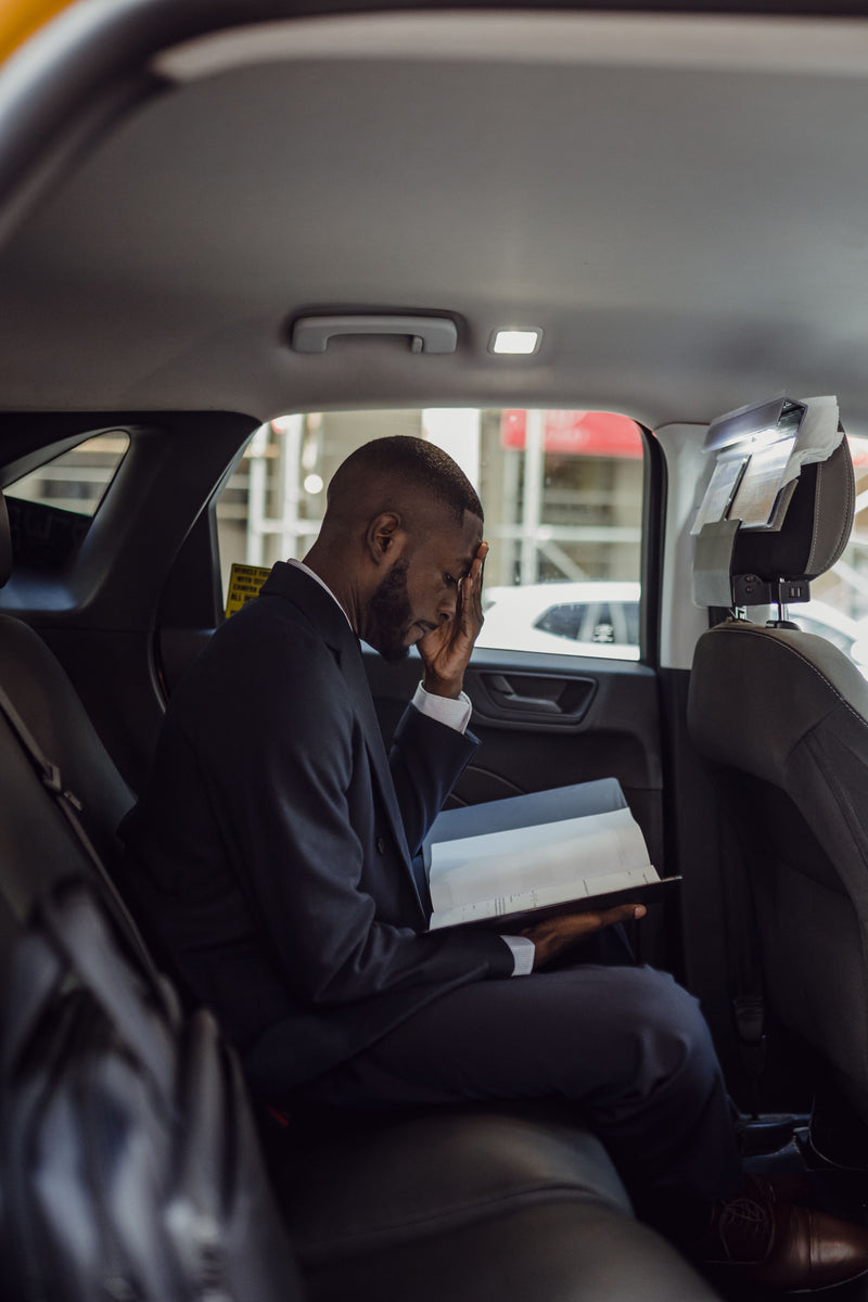 A man in a dark suit sits in the back seat of a car, holding an open book or tablet with his hand near his face.