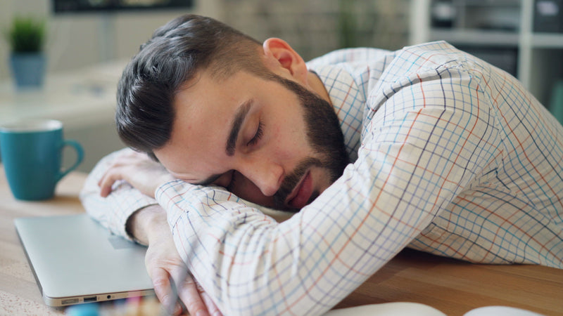 A man with a beard and short dark hair rests his head on his arms on a wooden desk, wearing a white long-sleeve button-up shirt with a subtle blue and orange grid pattern. A blue mug and a closed silver laptop are on the desk next to him, with a softly blurred indoor background.