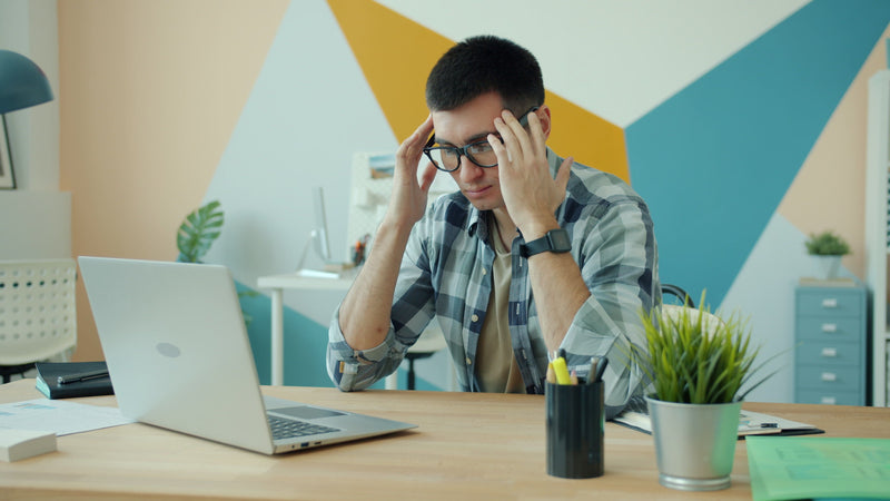 A man wearing glasses sits at a wooden desk in an office, holding his head in frustration with a laptop open in front of him. The background features a colorful geometric wall design in blue, yellow, and beige.