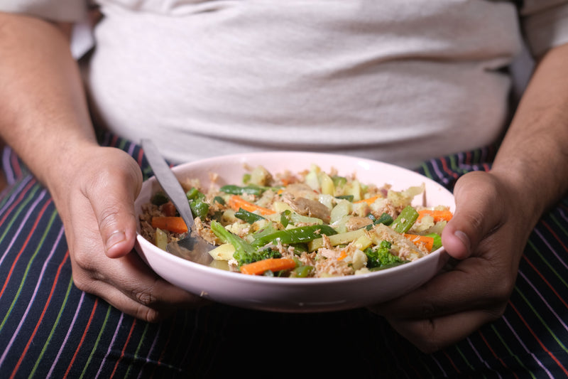 A close-up of a person holding a white bowl filled with a colorful vegetable and grain salad, with a spoon inside. The person is wearing a white shirt and a striped apron with green, red, blue, and black stripes.