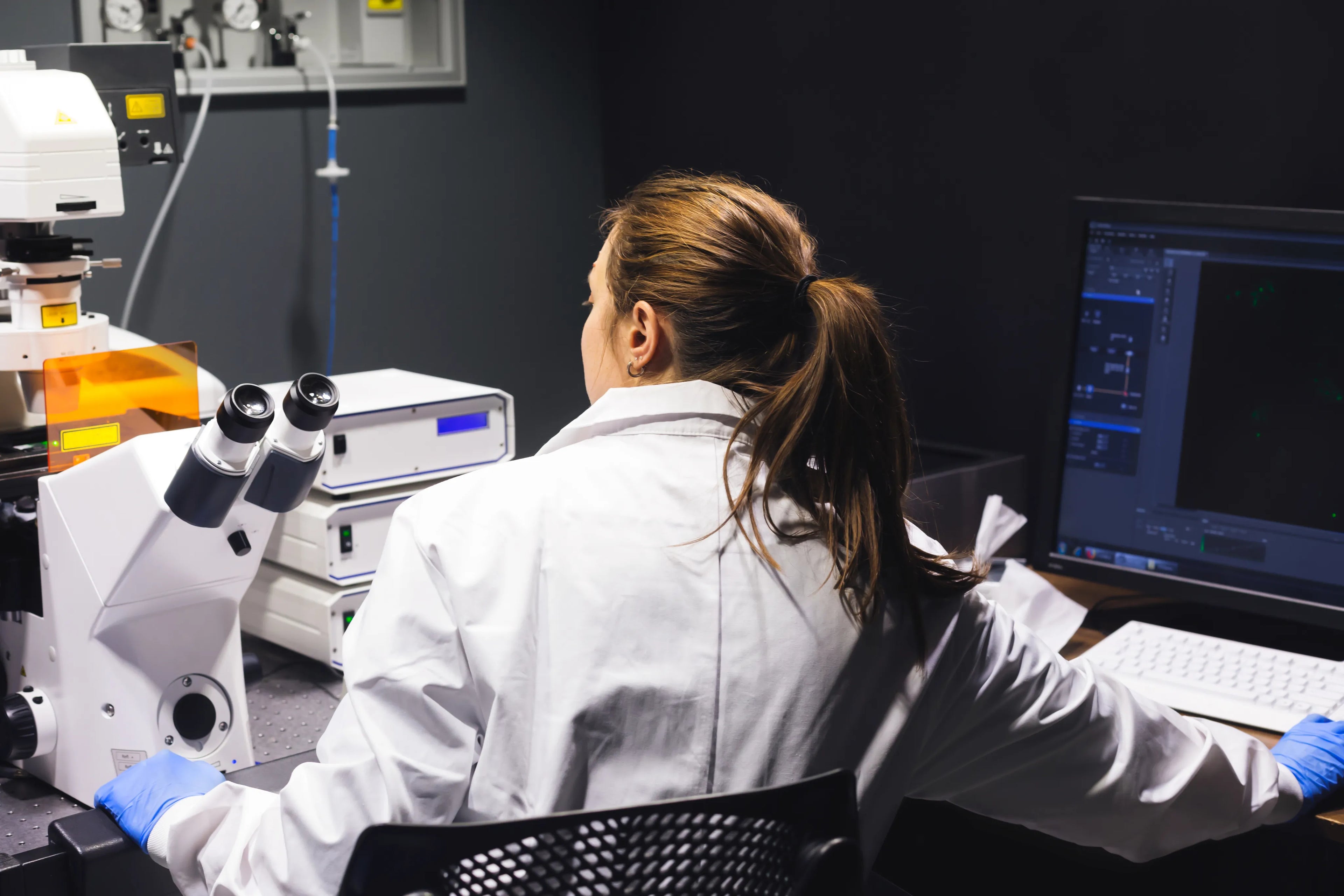 Person in a lab coat working at a computer with laboratory equipment in the background